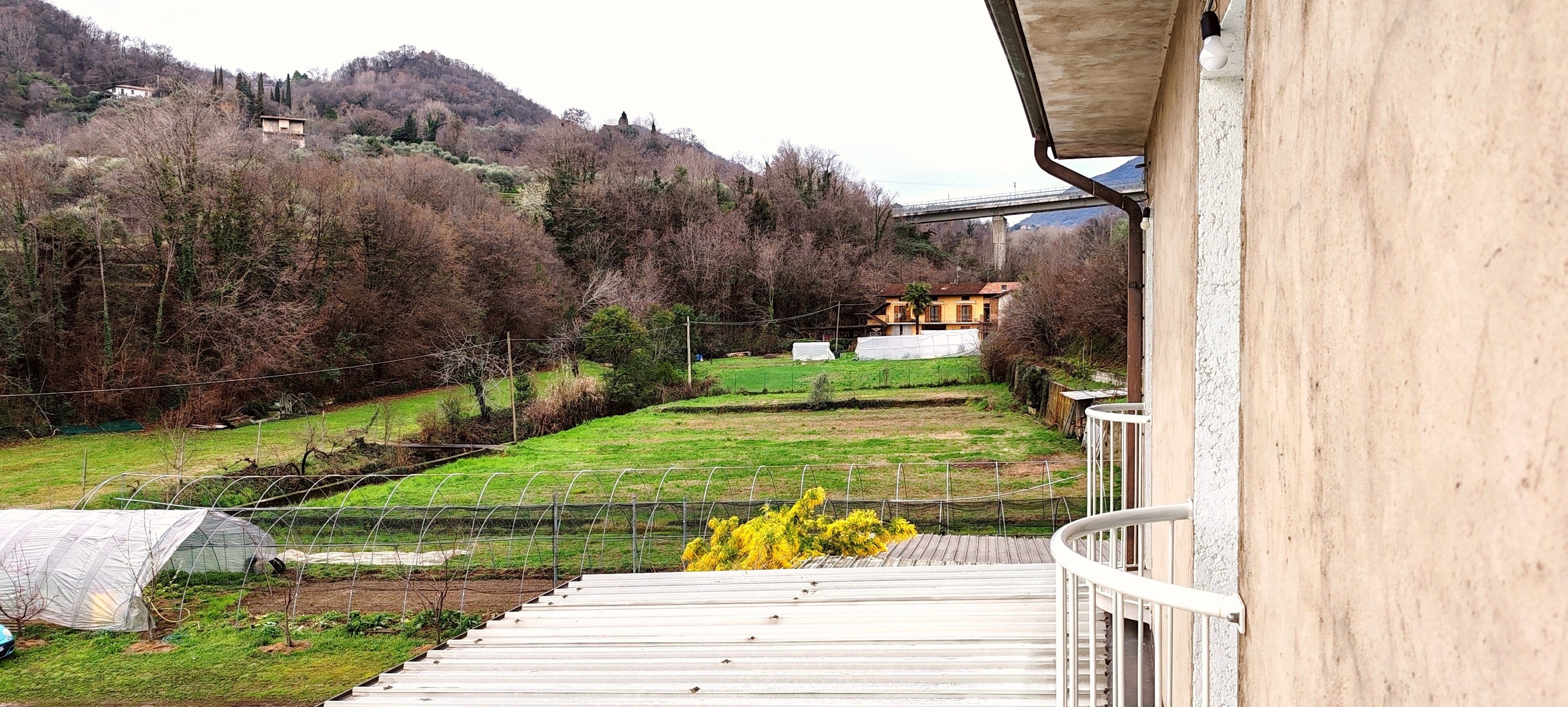 A balcony with a view of a green field, a yellow bush, and a distant house on a hill.