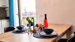 Ein Essbereich mit Holztisch, gedeckt für zwei Personen, einer Blumenvase, einer Weinflasche und Blick ins Freie durch ein Fenster.