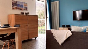 Ein Wohnzimmer mit Holzschrank, einem an der Wand befestigten Fernseher, einer Couch mit Kissen und Blick ins Freie durch eine Glastür.