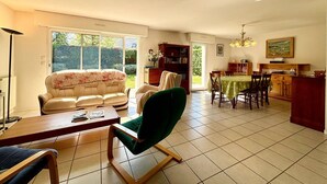 A spacious living room with a sofa, a green armchair, a dining table with chairs, a kitchen area, and a bookshelf. - Carnac: 5-room house with garden, 7 people, WiFi, pets allowed (Carnac)