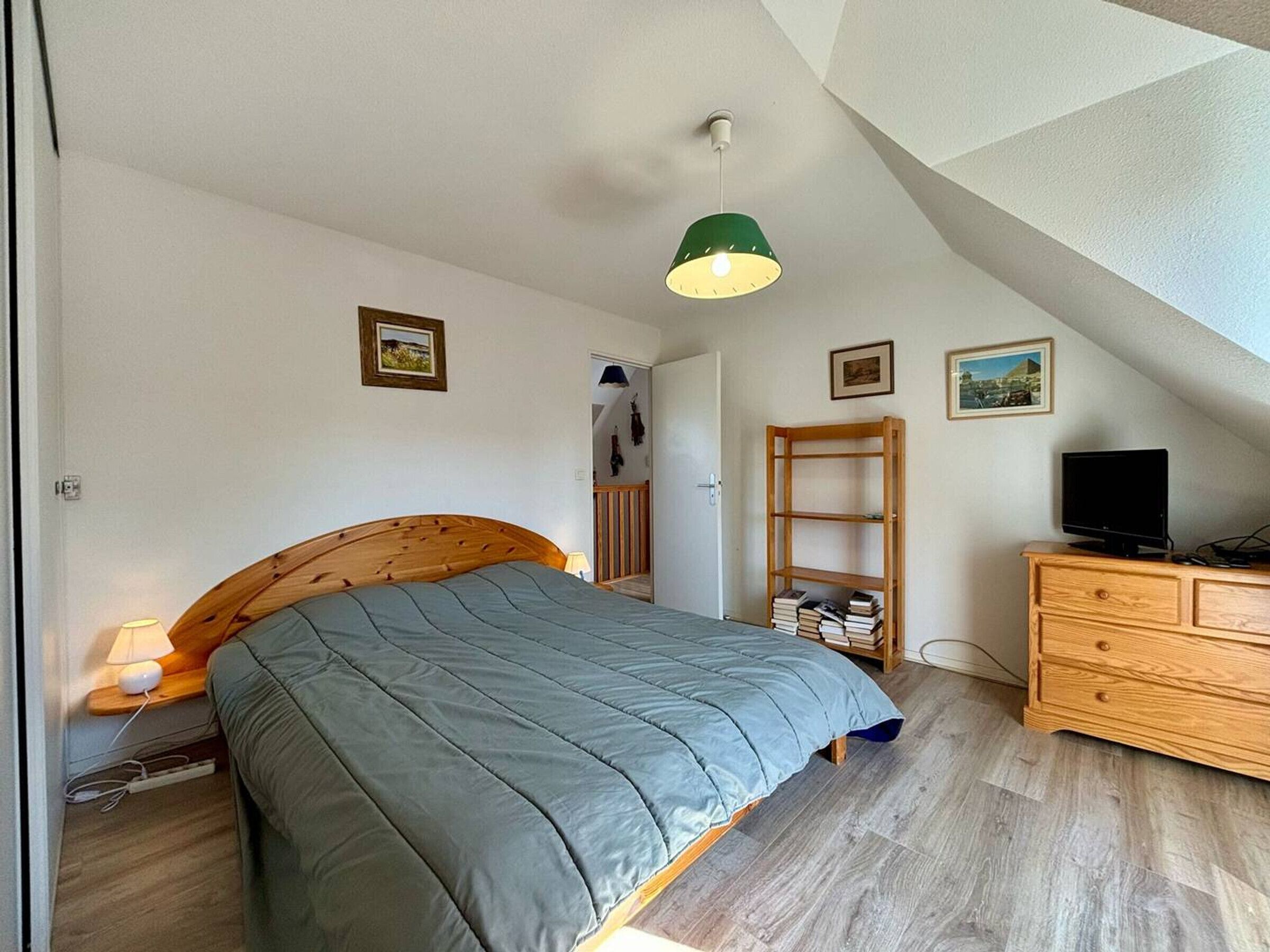 Un dormitorio con una cama de madera, una lámpara colgante verde, un aparador de madera y un televisor.