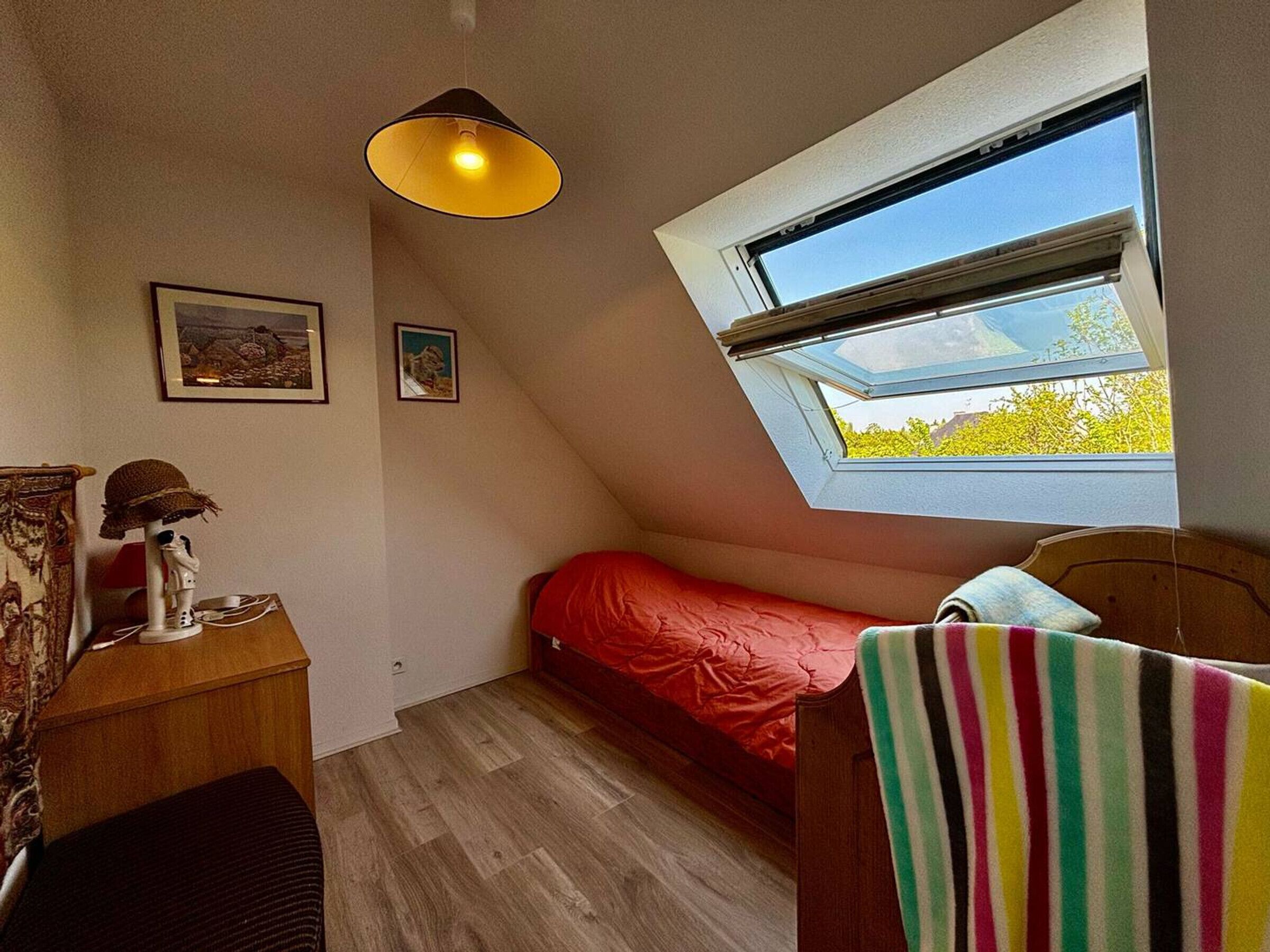 Et lille, hyggeligt værelse med skråvægge, en enkeltseng med et rødt tæppe, et trænatbord med en lampe og et vindue med udsigt til grønne områder.