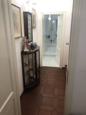 Un corridoio con pavimento piastrellato, una porta in vetro che conduce a un bagno e un armadietto in legno con ante trasparenti.