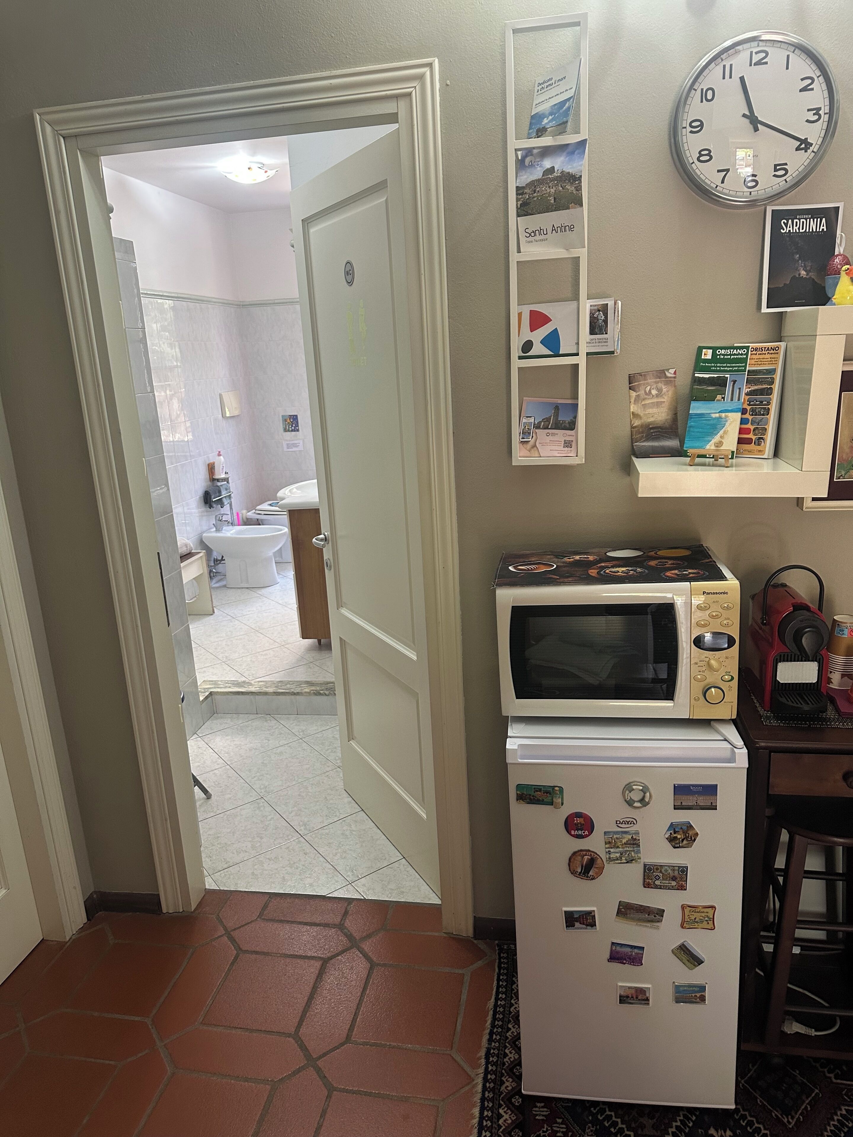 A kitchen with a microwave and refrigerator, a clock on the wall, and a door leading to another room.