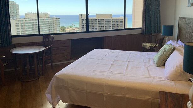 Camera d'albergo con un'ampia finestra che si affaccia sull'oceano e sugli edifici. Si vede un letto con i cuscini, una poltrona, un tavolino e una scrivania.
