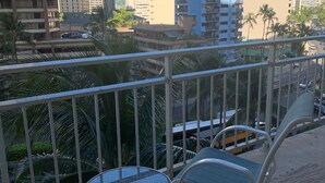 Un balcón con dos sillas y una pequeña mesa, con vista a un paisaje urbano de edificios altos y palmeras.
