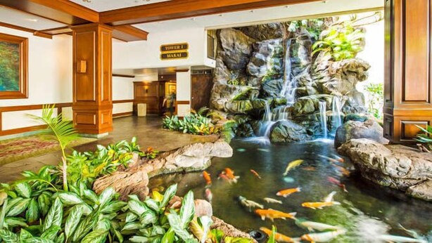 A hotel lobby with a koi pond, a waterfall, and a sign indicating 'Tower 1' and 'Makai'. - 29th Floor Waikiki Banyan, Full Kitchen and Free Parking (Honolulu)