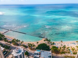 A beach with clear turquoise waters, a pier, and a parking area with cars.