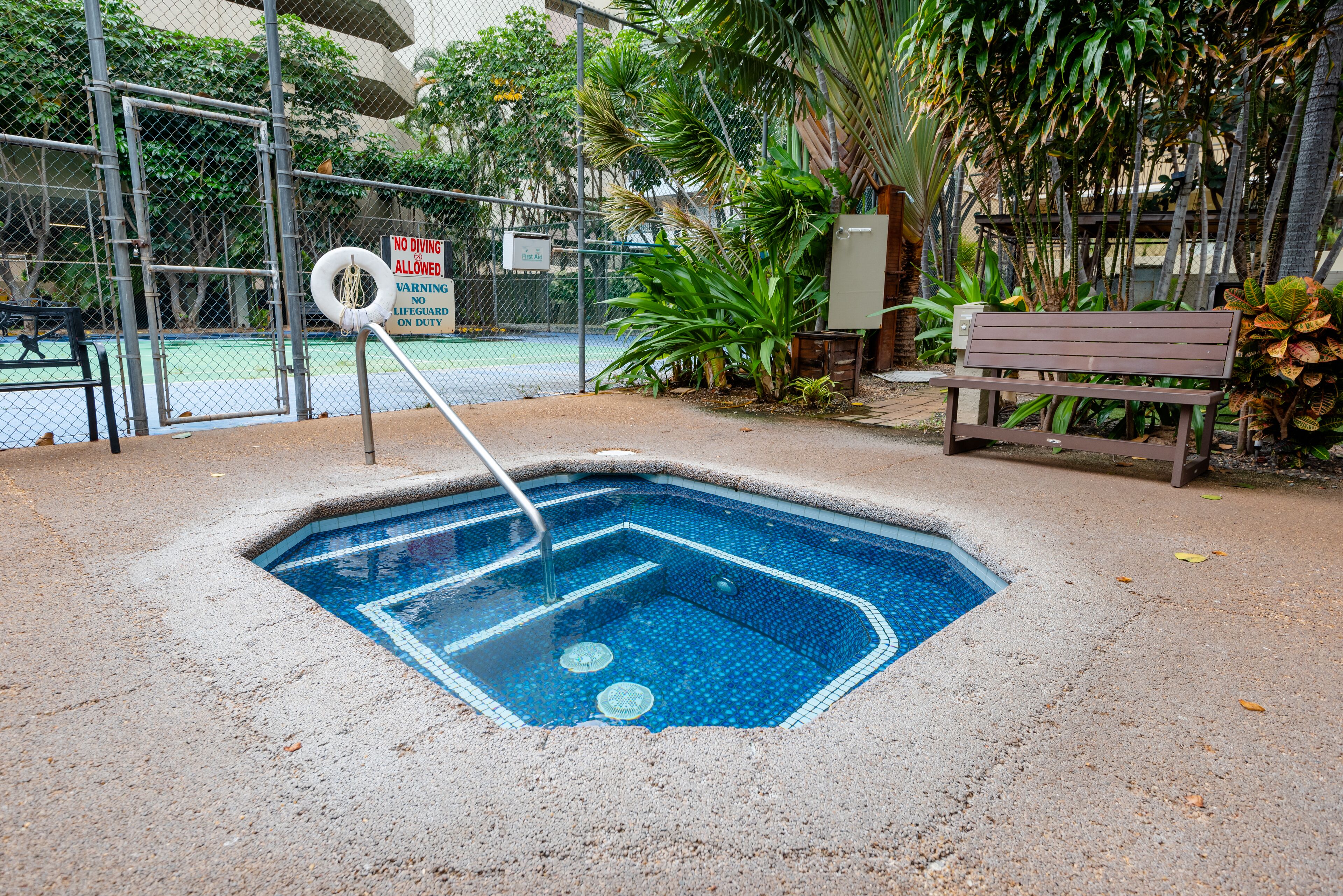 A small, square hot tub with a metal ladder, surrounded by a concrete edge and a fence.