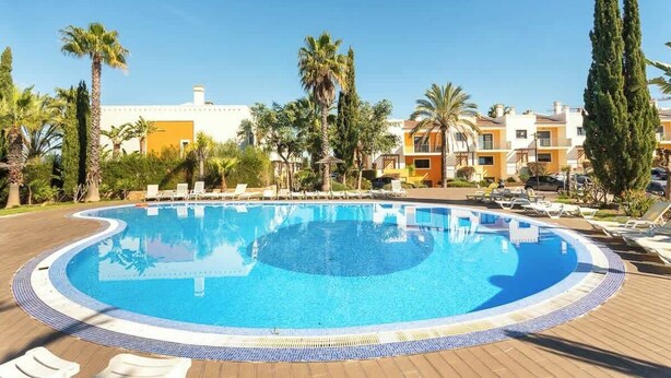 A swimming pool surrounded by lounge chairs and palm trees, with buildings in the background.
