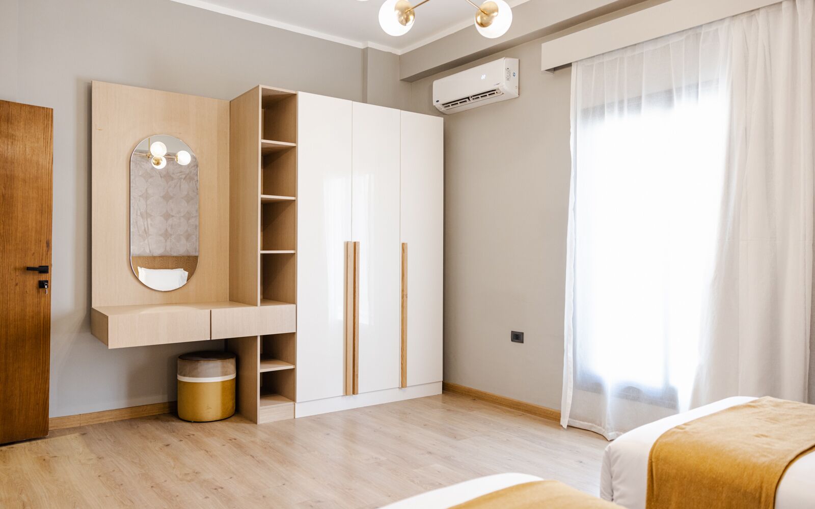 Un dormitorio moderno con un armario de madera, un espejo, un cubo de basura pequeño y una cama con manta blanca y color mostaza.