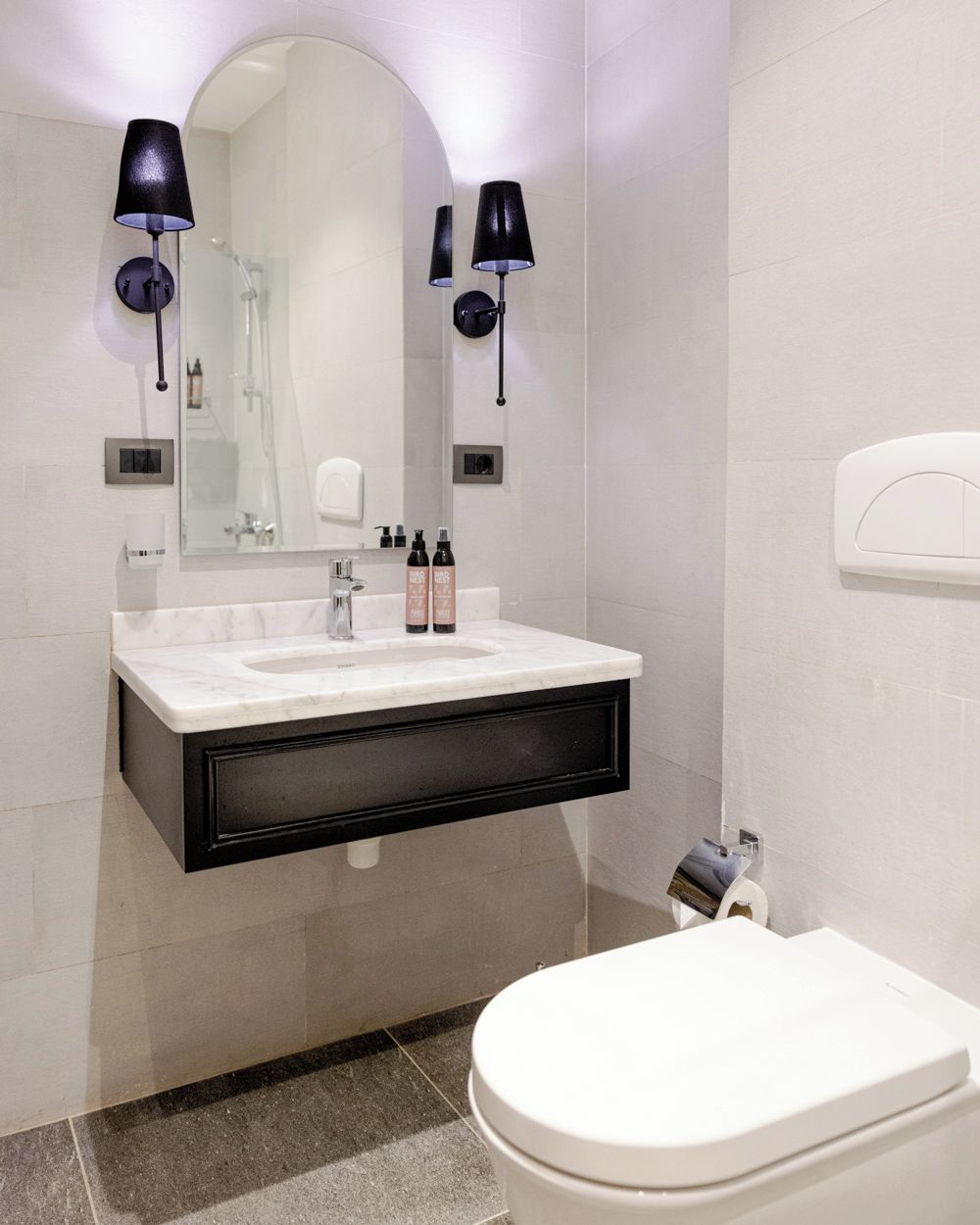 A modern bathroom with a white sink, two black wall-mounted lamps, a mirror, and a toilet.
