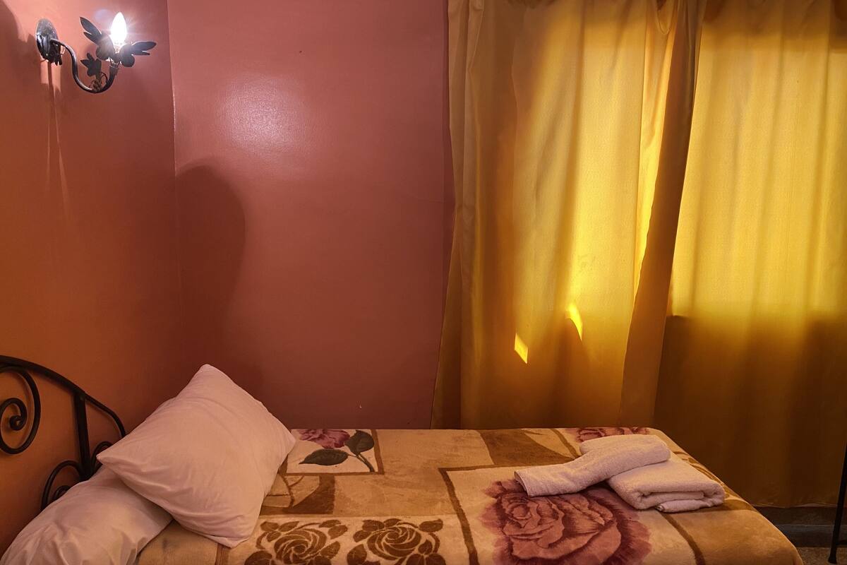 A bedroom with a bed, pillows, a floral bedspread, a wall-mounted light fixture, and a window with curtains.