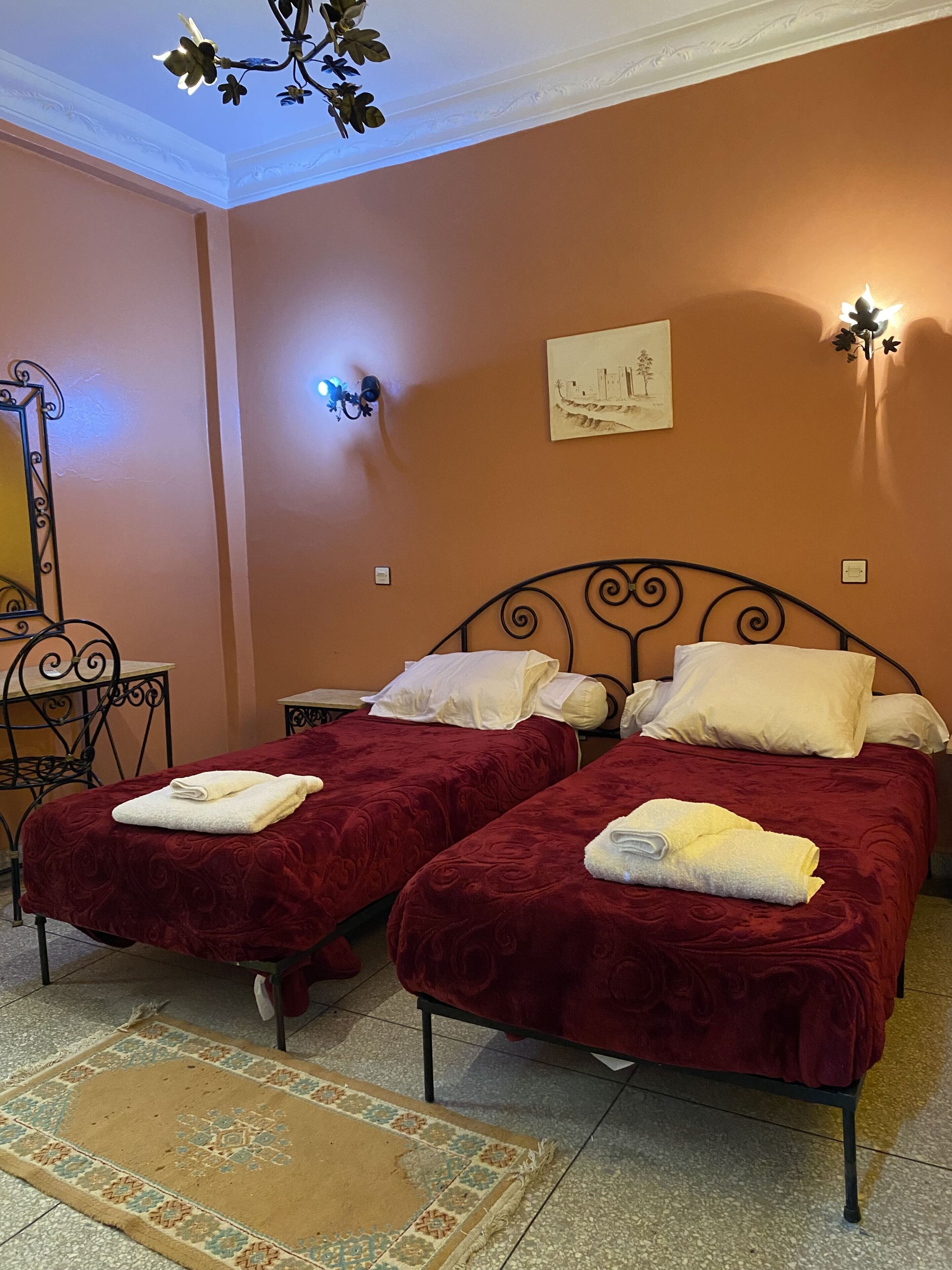 Habitación con dos camas, cada una con cubrecama rojo y almohadas blancas, una mesita con una silla y un cuadro colgado en la pared.