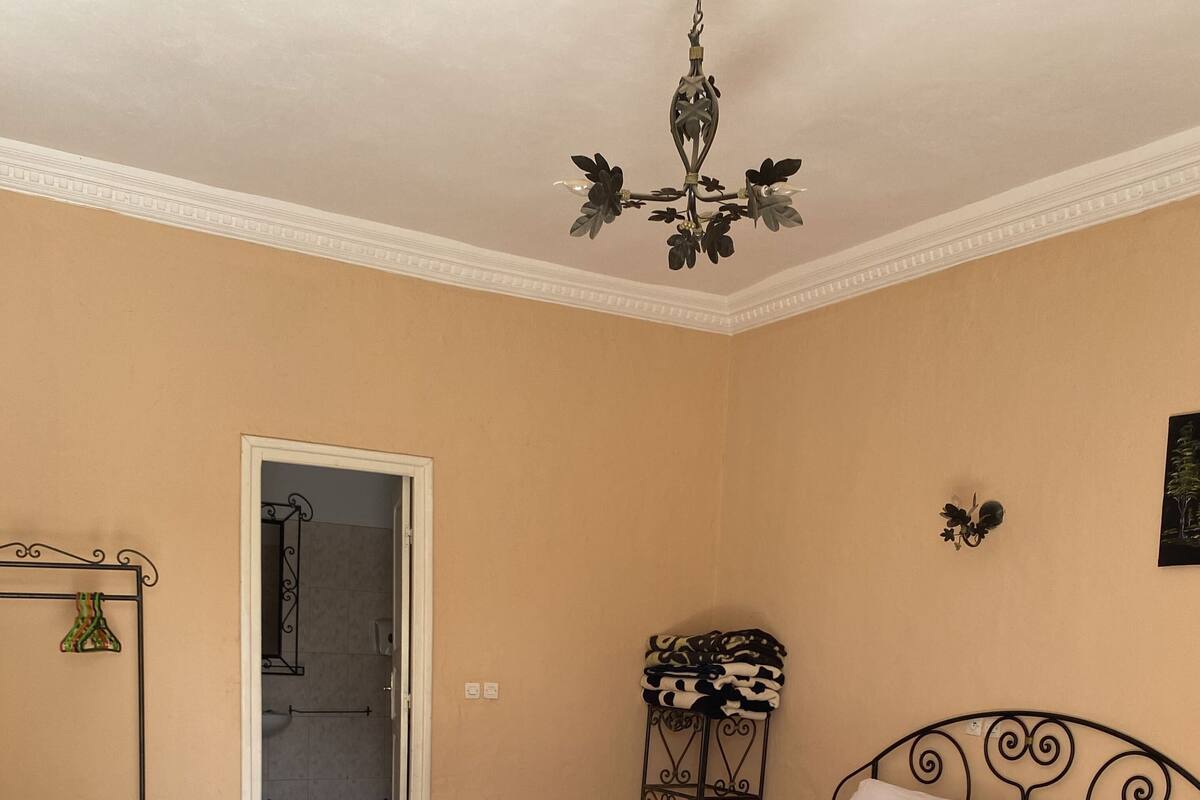 A bedroom with a bed, a bench, a small table, a hanging light fixture, and a door leading to a bathroom.