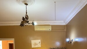 A bedroom with a bed, a ceiling fan, an air conditioning unit, a wall-mounted light fixture, and a door leading to another room.