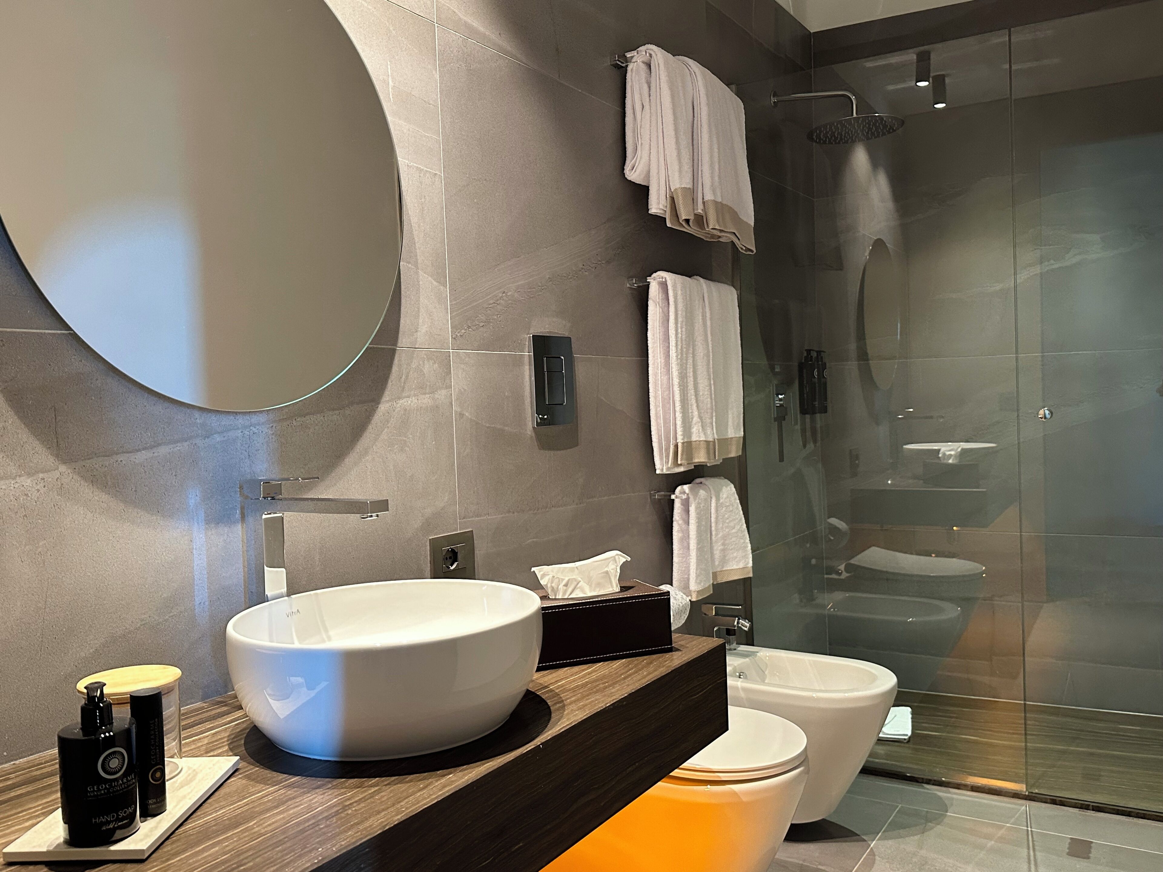 Un baño moderno con un espejo redondo grande, un lavabo sobre encimera, inodoro y cabina de ducha con paredes de vidrio.