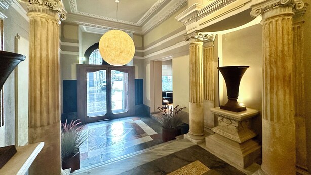A grand hallway with classical architecture, featuring columns, a large chandelier, and a view into the adjacent room. - Palazzo Liberty (Palermo)