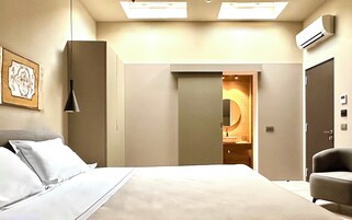 A modern hotel room with a large bed, a desk, and a chair. There is a door leading to a bathroom with a round mirror and a toilet. The room has a ceiling light and an air conditioning unit.