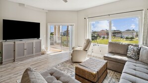 A spacious living room with a flat-screen TV, a ceiling fan, a large window with a view of the neighborhood, and a balcony with a chair. - Ocean Park 114 - Hosted by Moore AI Rentals (Fernandina Beach)