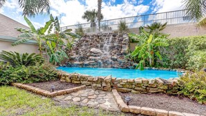 A backyard with a swimming pool, a stone waterfall, and tropical plants. - Ocean Park 114 - Hosted by Moore AI Rentals (Fernandina Beach)