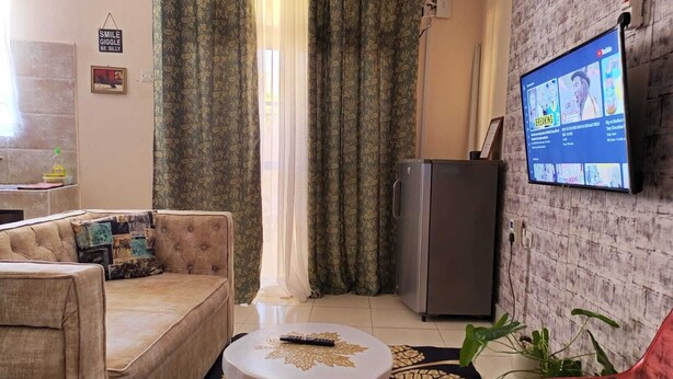 A living room with a sofa, a small table, a television mounted on the wall, a refrigerator, and a plant. - Lux Suites La Vida Studio Apartment (Mombasa)
