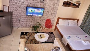 A hotel room with a brick-patterned wall, a bed with white linens, a wooden headboard, a red chair, a small white table with a gold leaf design, and a television mounted on the wall. - Lux Suites La Vida Studio Apartment (Mombasa)