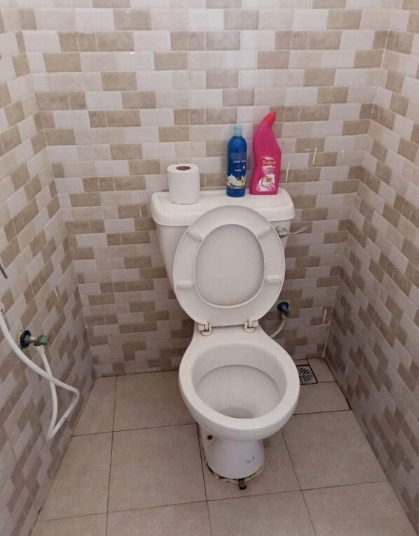 Un baño pequeño con azulejos, un inodoro, un rollo de papel higiénico y dos botellas de artículos de tocador sobre el tanque.