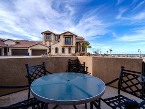A patio with a round glass table and black metal chairs, overlooking a coastal area with a large house and beach in the background.