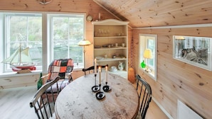 Interior acogedor de una cabaña, con paredes de madera, una mesa de comedor con velas, una silla, una ventana con vistas a follaje y una estantería con vajilla.