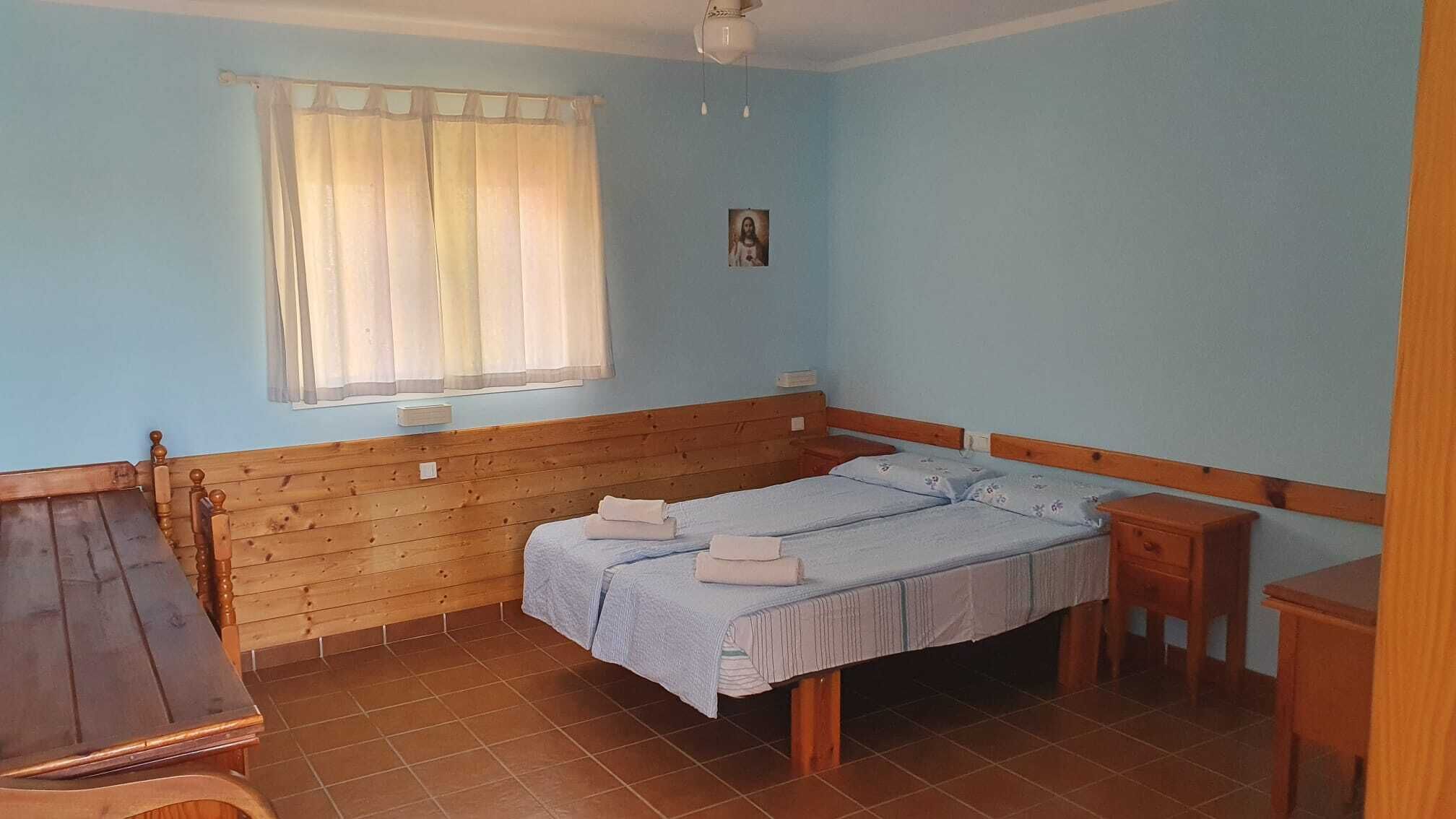 A small, single-room accommodation with a wooden bed, a small table, a chair, and a window with curtains.