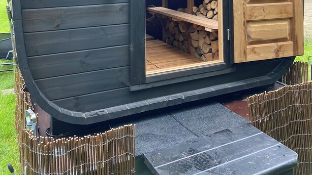 Une petite cabane en bois, dont la porte est ouverte, laissant apercevoir une étagère en bois et une pile de bois de chauffage à l’intérieur.