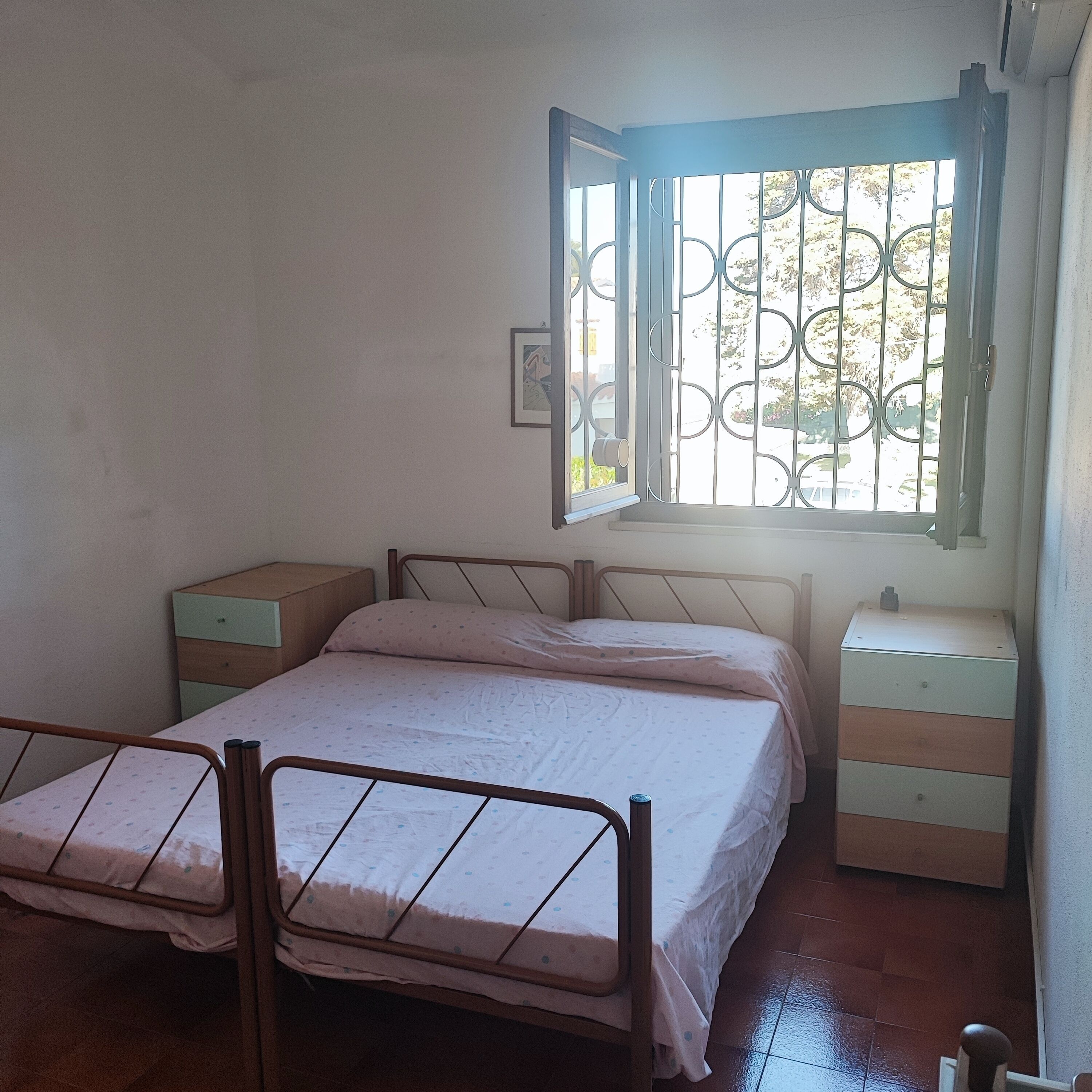 Een slaapkamer met een enkel bed, nachtkastje, dressoir en een raam met metalen tralies.