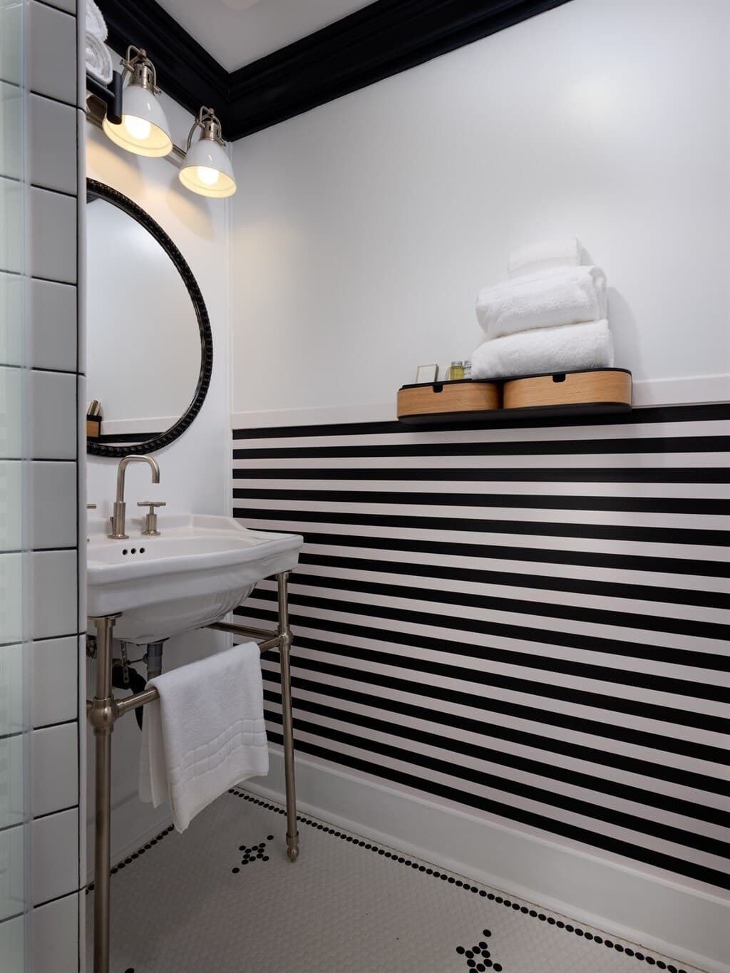 A bathroom with a white sink, a mirror, and two wall-mounted lights. There is a towel rack with towels and a wooden tray with toiletries.