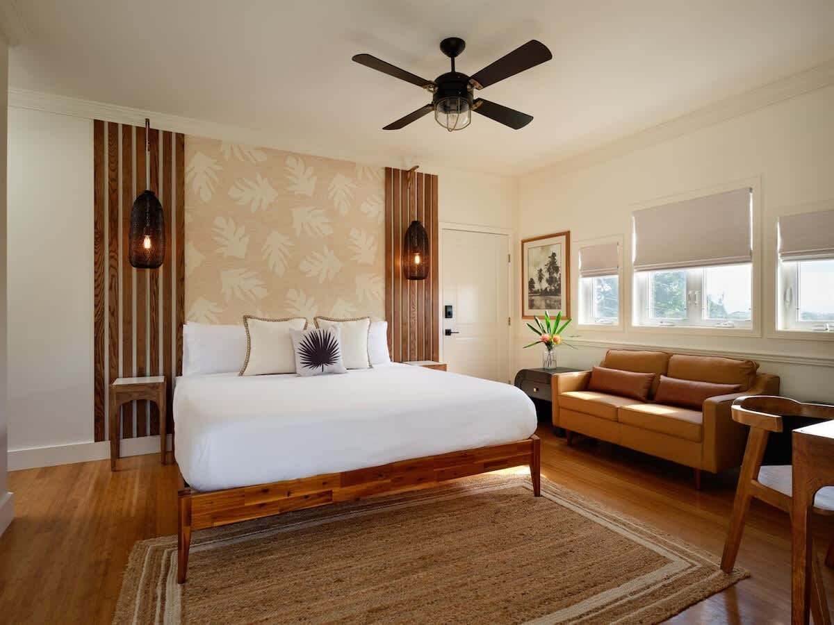 A bedroom with a bed, a wooden headboard, a ceiling fan, a nightstand, a bedside table, a lamp, a window with blinds, a sofa, a chair, and a rug.