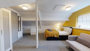 A bedroom with a bed, a bedside table, a lamp, a sofa, and a window with blinds.