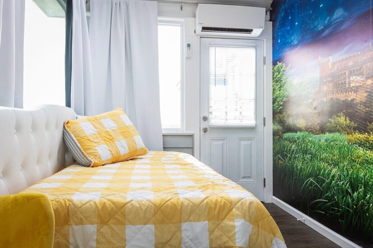 Ein Schlafzimmer mit kariertem Bettbezug, einem weißen, gepolsterten Kopfteil und einer Tür mit Blick auf eine grüne Landschaft.