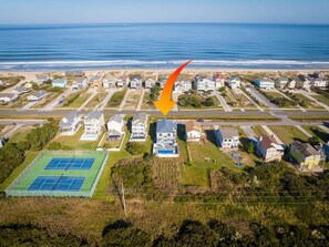 Vue aérienne d’un quartier résidentiel situé sur la côte, avec des maisons, un court de tennis et une plage en arrière-plan.