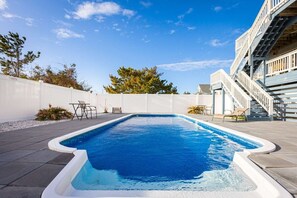 Une piscine rectangulaire bordée de blanc, entourée d’une terrasse dallée avec des chaises et des tables.