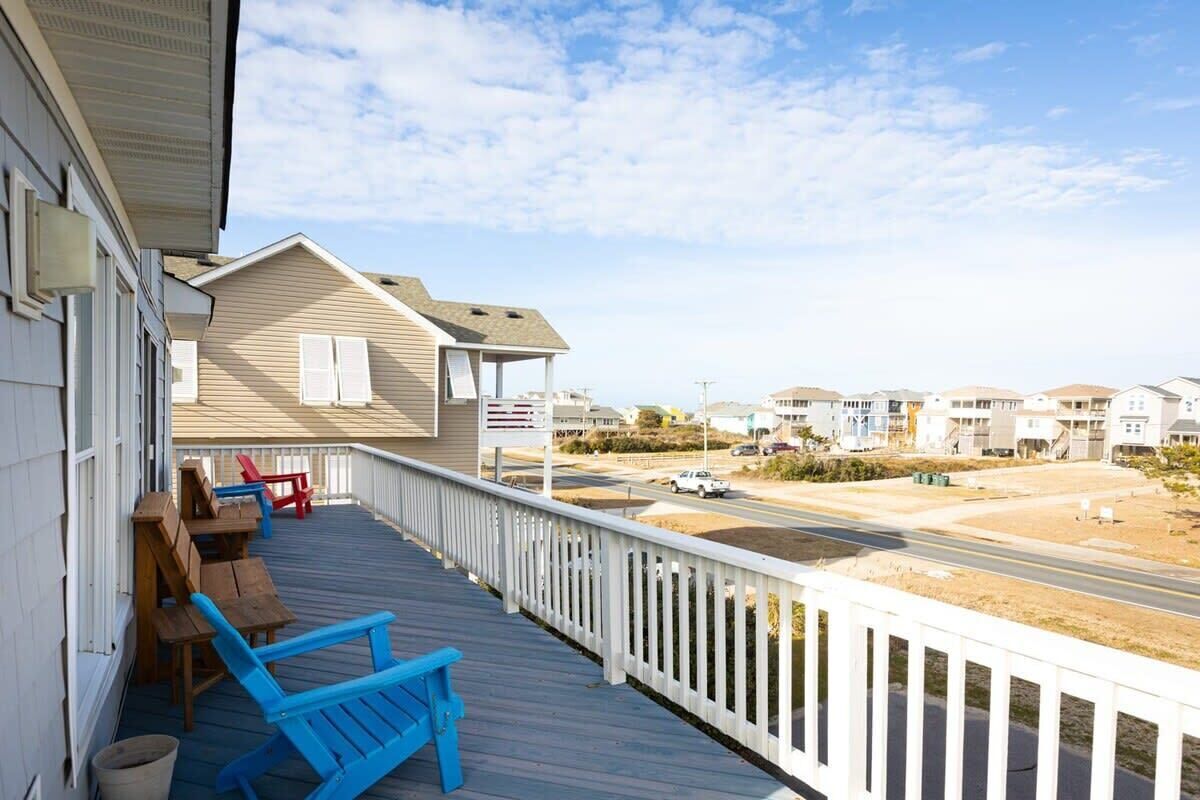 Une terrasse en bois avec des chaises bleues et une balustrade blanche, donnant sur une route et des maisons.