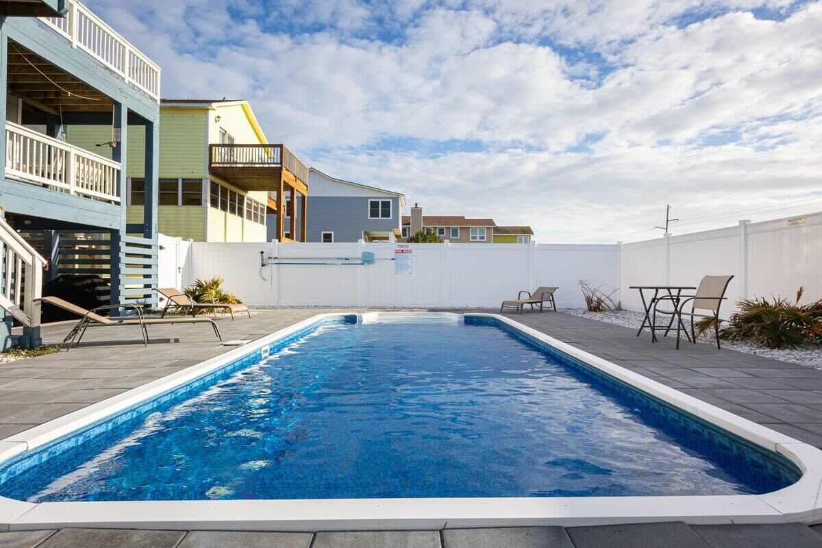 A rectangular swimming pool with a white border, surrounded by a patio area with chairs and a table.