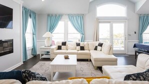 Amplia sala de estar con un sofá modular grande, una mesa de centro blanca y un ventilador de techo. Se observan ventanas amplias con cortinas azules, un televisor de pantalla plana montado en la pared y un balcón visible a través de las ventanas.