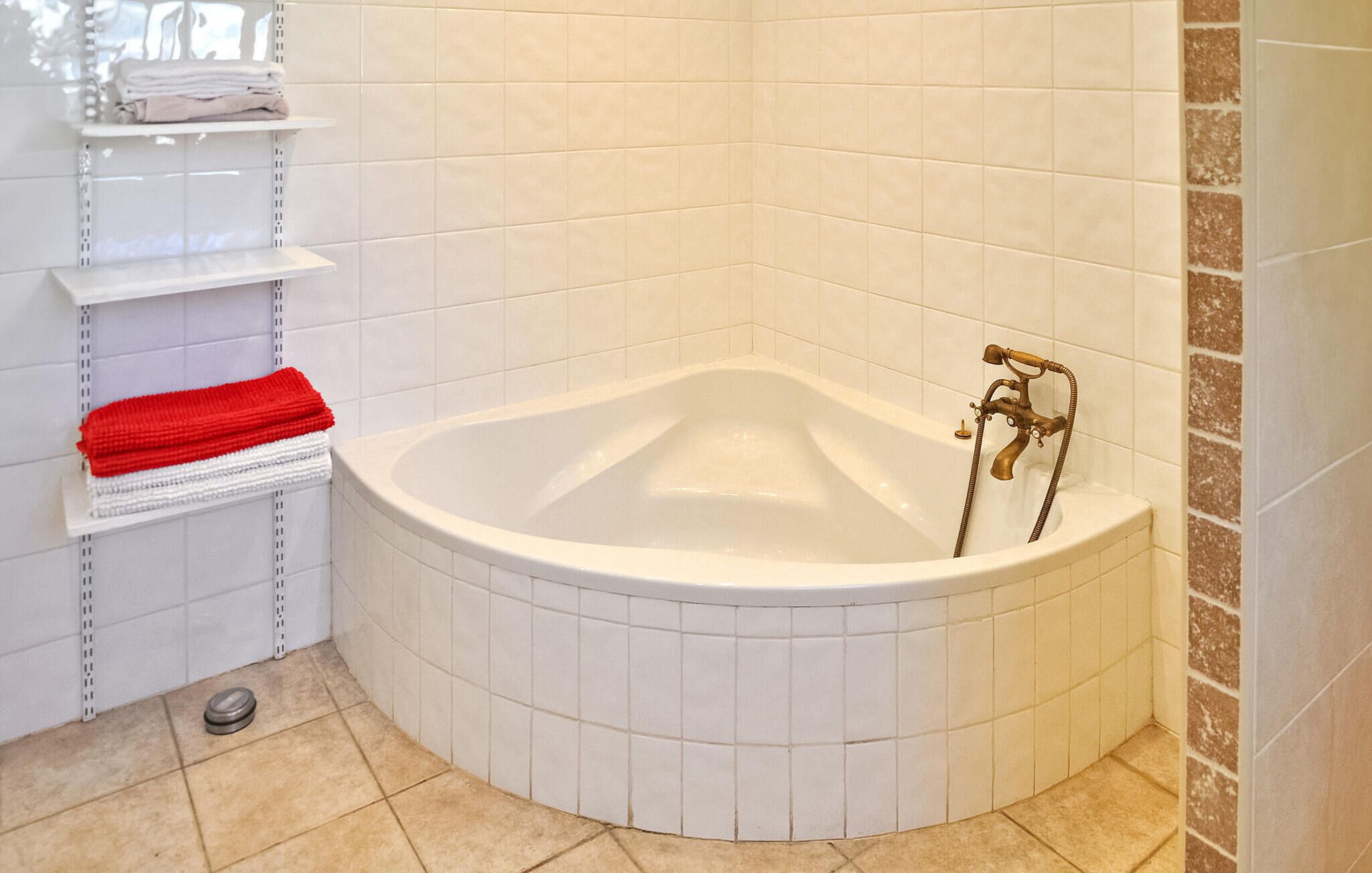 A corner bathroom with a freestanding bathtub, white tiled walls, a shelf with towels, and a metal floor drain.