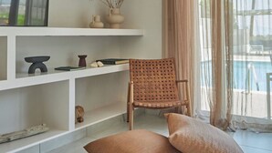 Een moderne woonkamer met een witte boekenkast, een rieten stoel en uitzicht op een zwembad door dunne gordijnen.