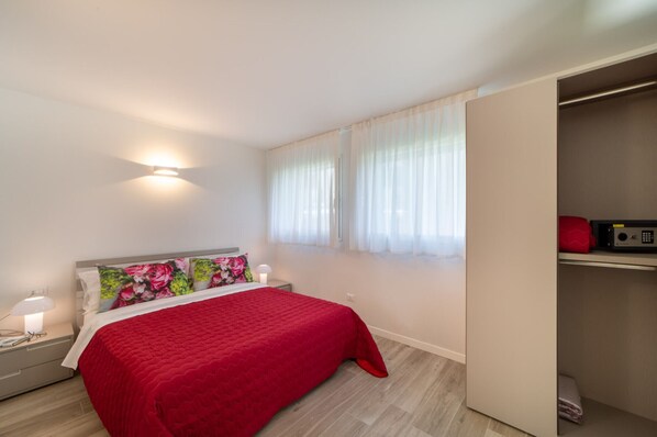 Un dormitorio con cama, mesita de noche, lámpara, ventana con cortinas traslúcidas y armario.