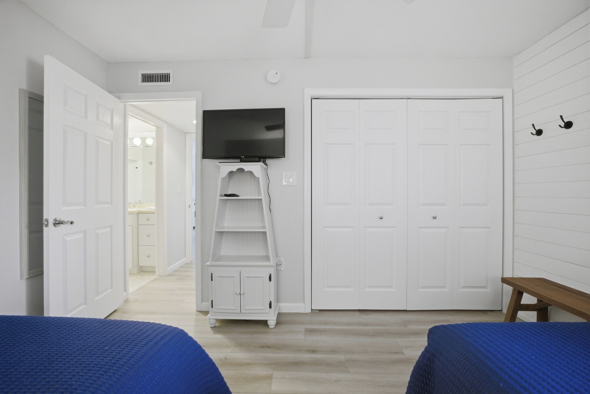 Quarto com duas camas, uma televisão em uma prateleira, um guarda-roupa e um banco.