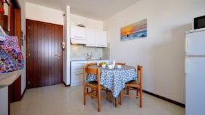 Een kleine keuken met een eettafel gedekt voor twee personen, een koelkast, en een schilderij van een zonsondergang aan de muur.