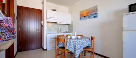 A compact kitchen with a dining table set for two, a refrigerator, and a painting of a sunset on the wall.
