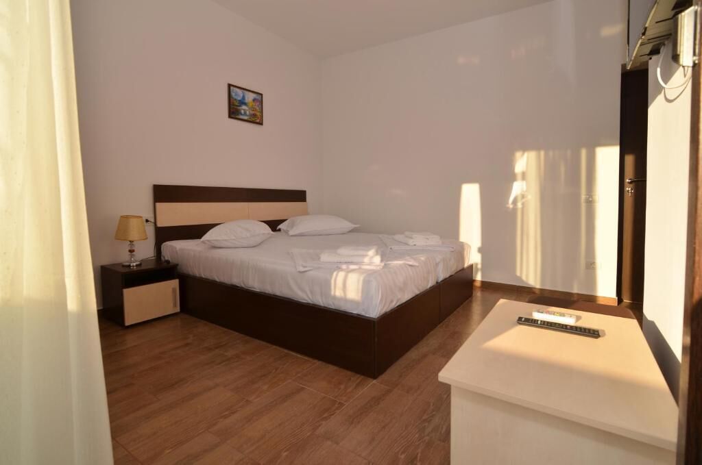 A neatly made bed in a hotel room with a wooden floor, a bedside table with a lamp, and a remote control on a small table.