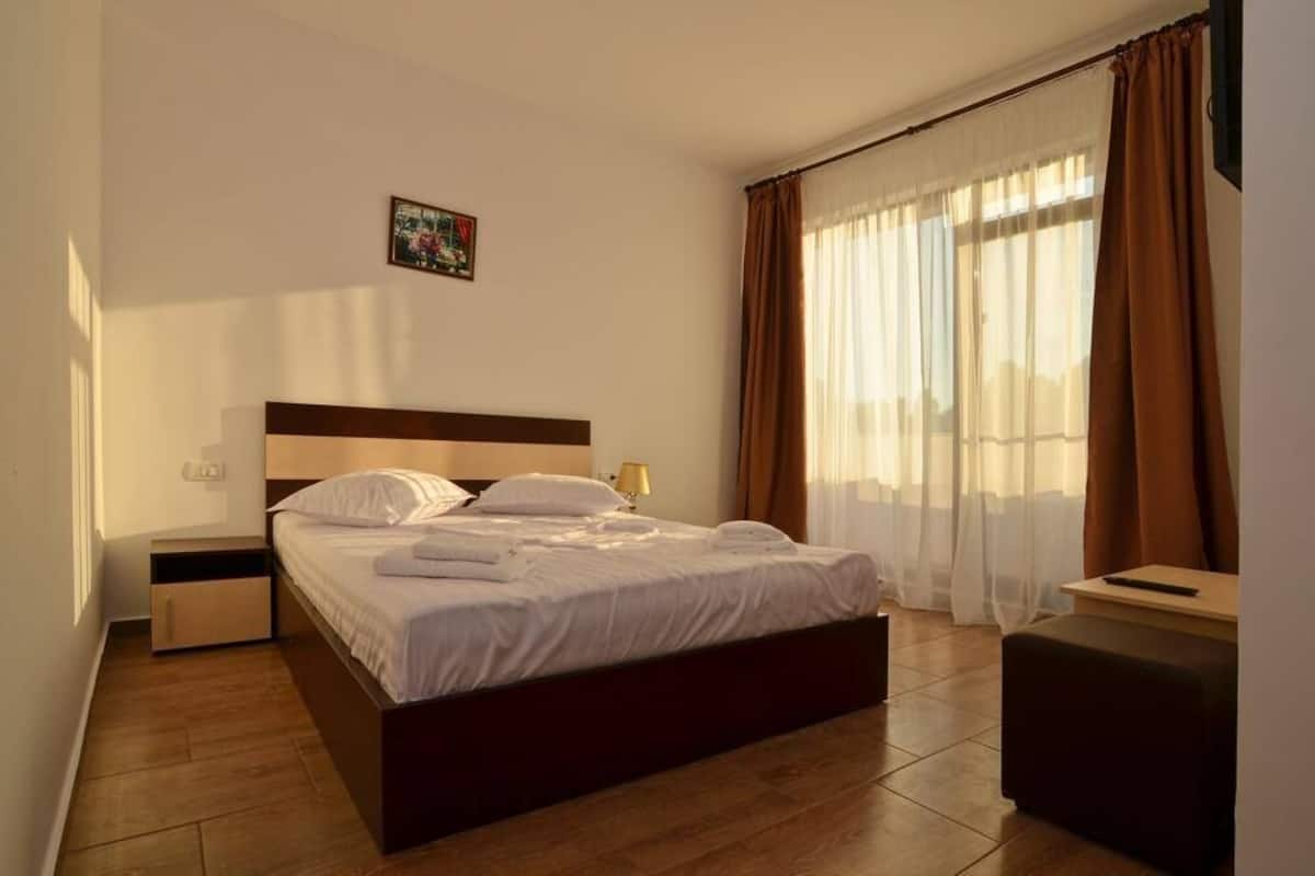 A neatly made bed with white linens and pillows, a bedside table with a lamp, a small drawer, and a window with sheer curtains and a brown drape.
